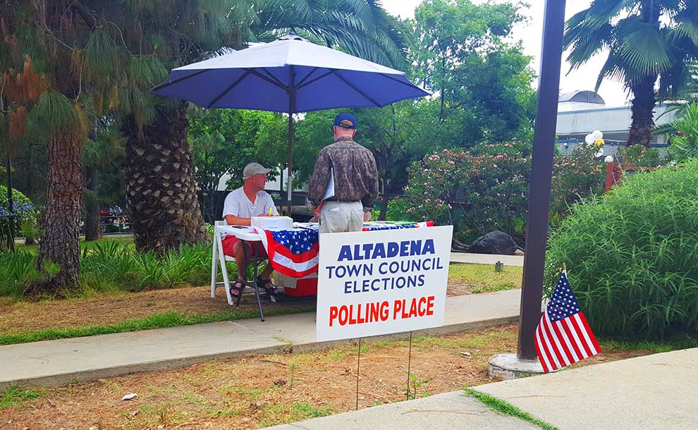 A man wearing a white hat under a blue umbrella talking to a voter in a brown shirt and a sign announcing the elections