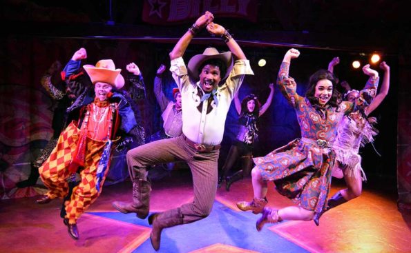 An African American coboy jumping in the air with a femae dressed in purple outfit behind him and a Cowboy clown to his left, all jumping in the air smiling