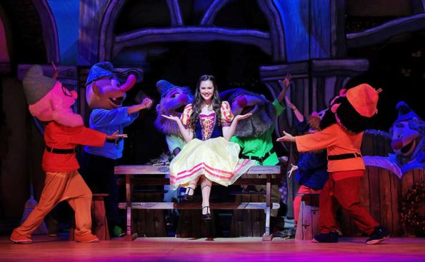 A girl dressed in white skirt and a blue vest surrounded by seven dwarfs