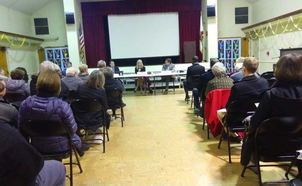 three candidates sitting behind a white table and aidience members listening