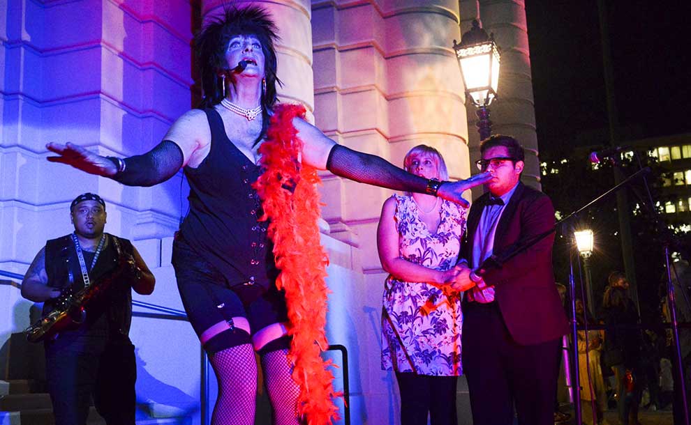 A drag person in a black dress and a red scarf singing and two people looking in awe