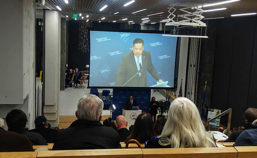 A man speaking on a podium with a huge screen behind him
