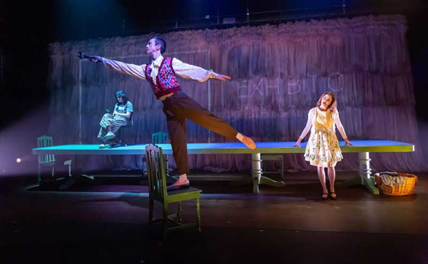 one actor on a table, another is balancing on a chair and a woman looking at him