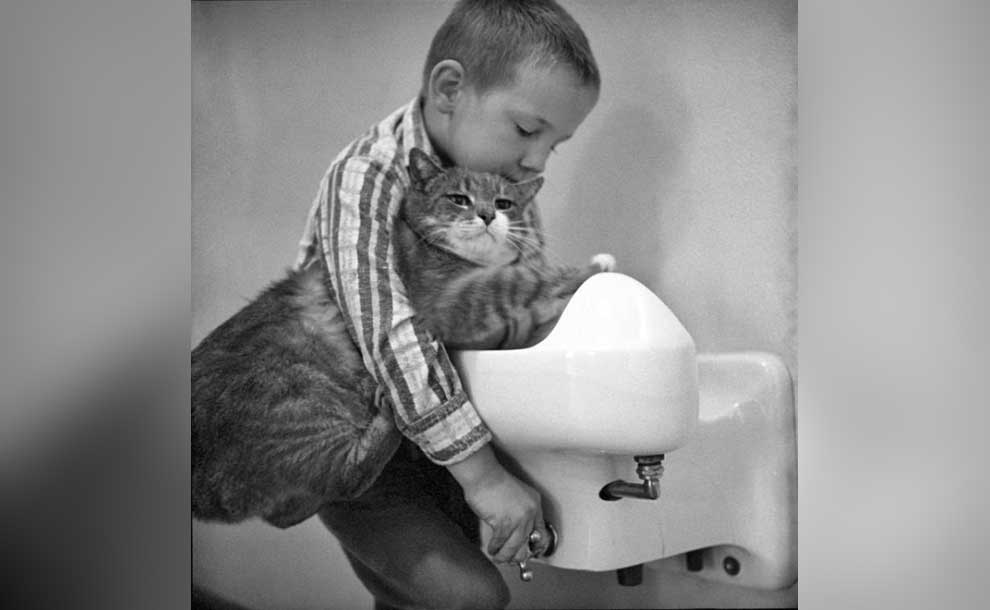 a kid attempting to drink from a fountain with a cat on his lap