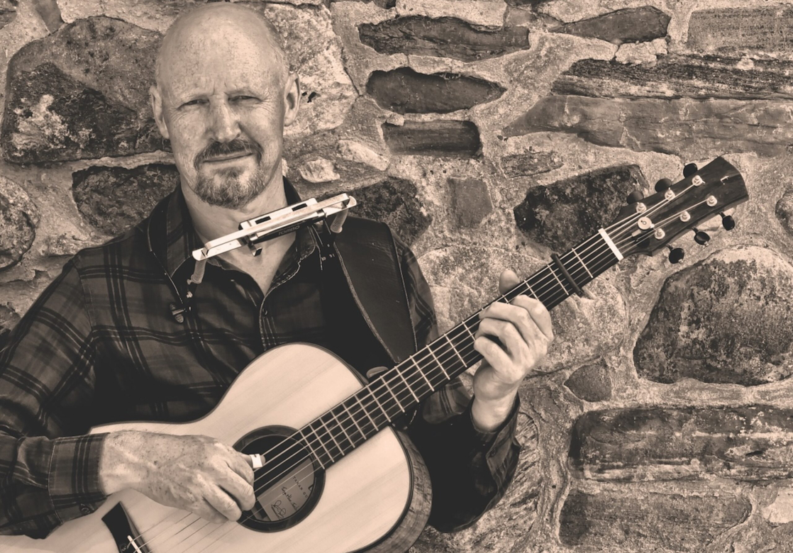 Singer/Songwriter Jim Malcolm holds a guitar and sports a harmonica in front of a stone wall
