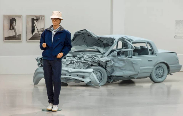 man with blue jack and white hat stands in front of grey sculpture of crashed car