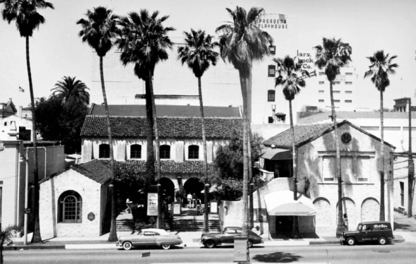 black and white photo of Spanish style three story building with tall palm trees and 1960s vehicles parked out front