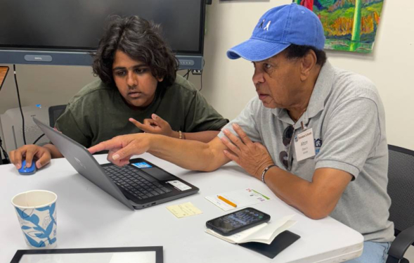 high school male student sits next to senior man in baseball cap as they both look at laptop screen while seated
