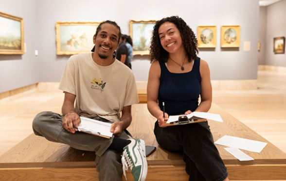 two smiling artists sit with sketch pads in a museum gallery