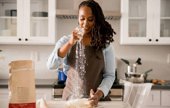 Black woman in kitchen smiles and adds flour to a bread loaf
