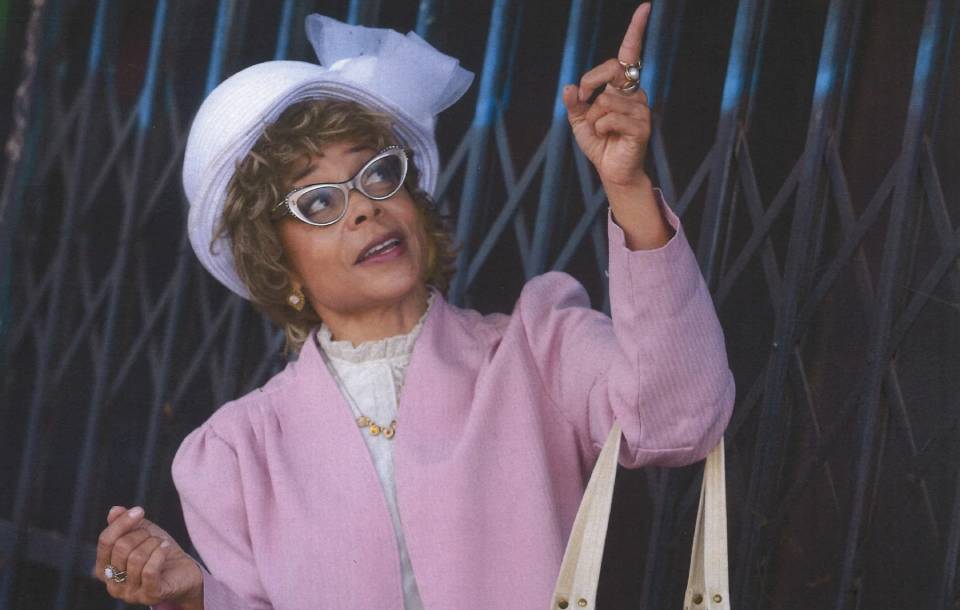 Black woman in light purple suit and white hat smiles and points to the ceiling