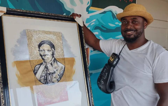Black man smiles at camera in white tshirt posed next to a framed portrait of Harriet Tubman