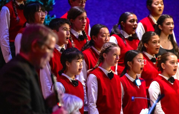 children in white shirts and red vests sing in front of twilight blue background on stage