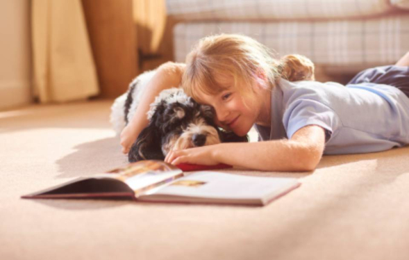 young girl lies next to a floppy eared dog on carpet with a book open in front of them