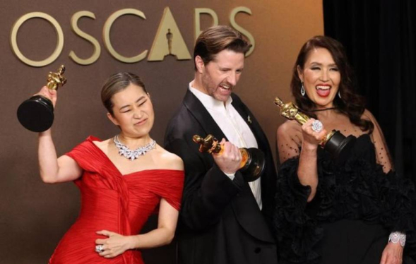 two women and a man, all in evening wear, cheer and hold golden statuettes aloft