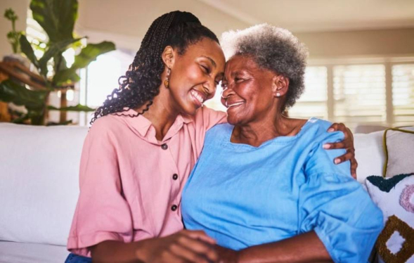 a younger woman embraces an older woman on a couch and both smile